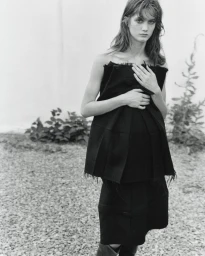Model in a black strapless pleated top and skirt standing outdoors on gravel against a light wall for The Courtyard project