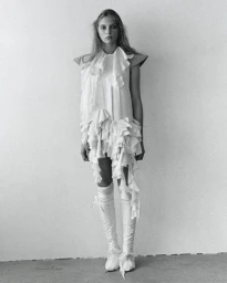 Model in a tiered ruffled white mini dress and knee-high white boots standing against a neutral studio backdrop.
