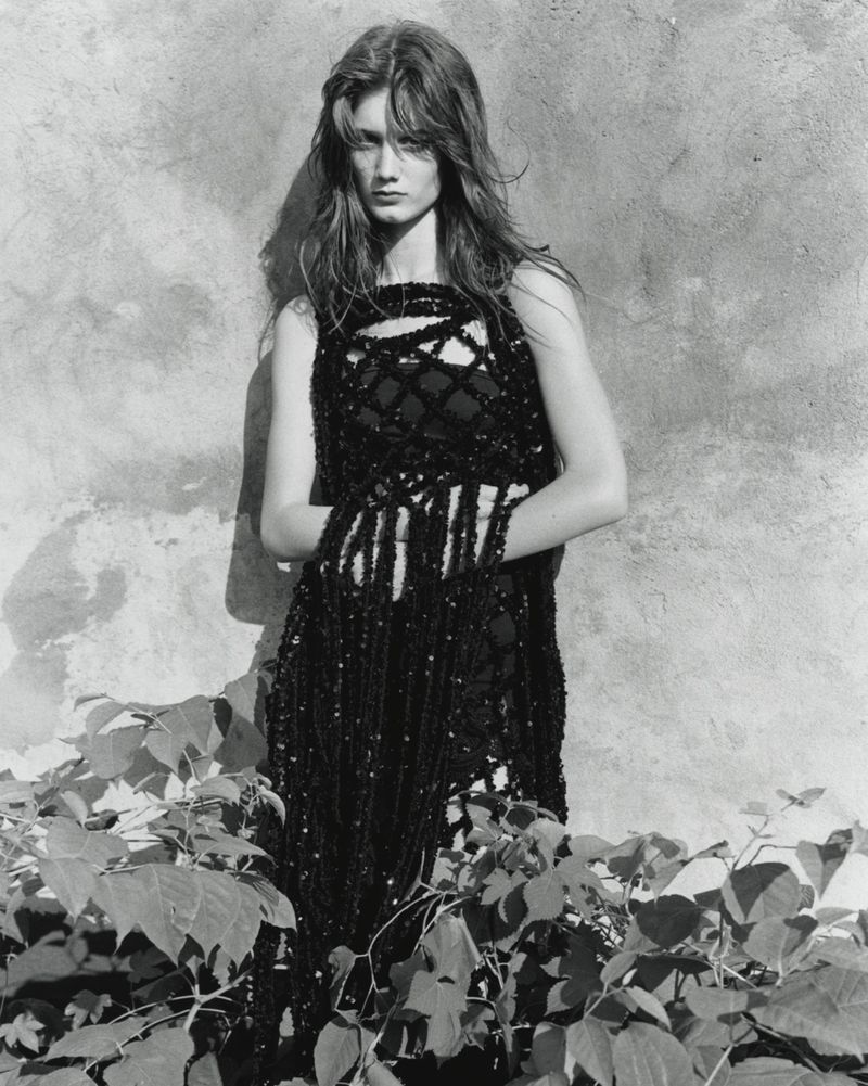 Model in black textured dress leaning against a weathered stone wall behind lush foliage in a black and white editorial portrait