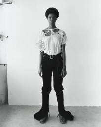 Monochrome full-length portrait of a Black female model wearing a distressed t-shirt and dark trousers against a plain white background.