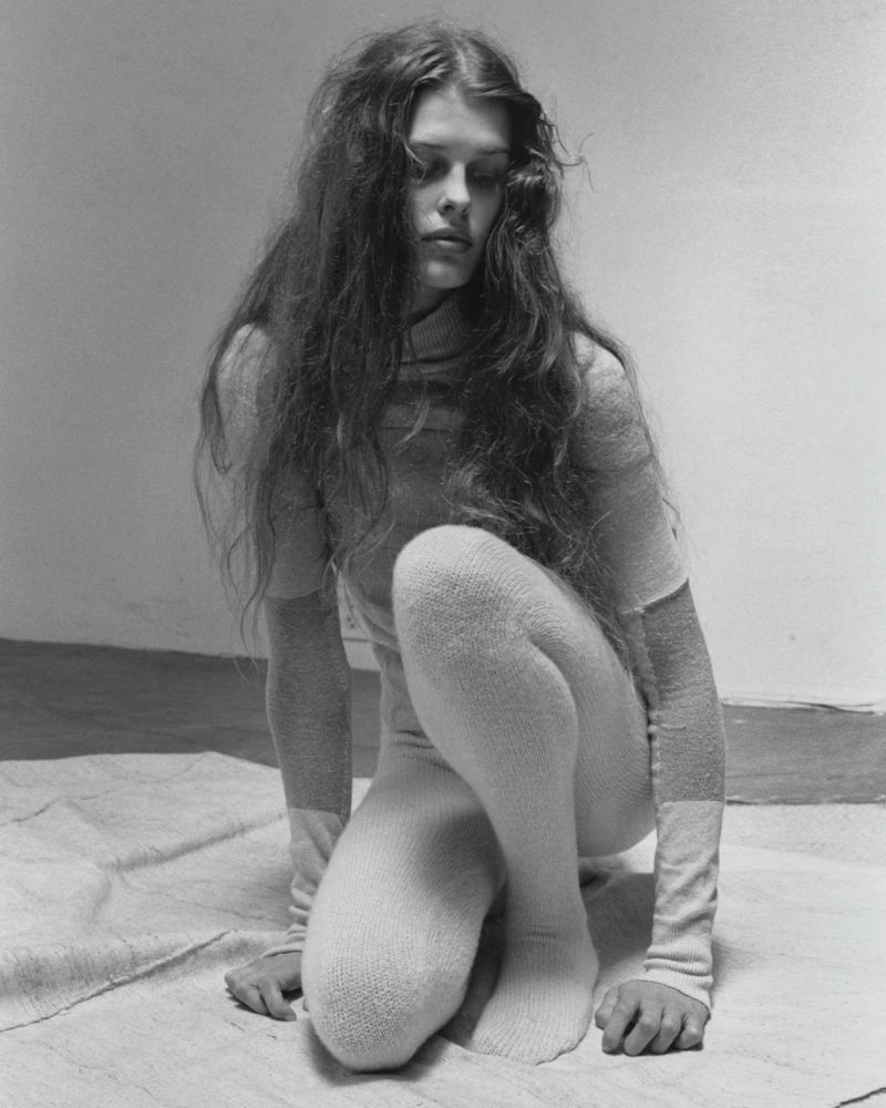 Black and white portrait of a female model crouching on a draped canvas surface in a minimalist studio.