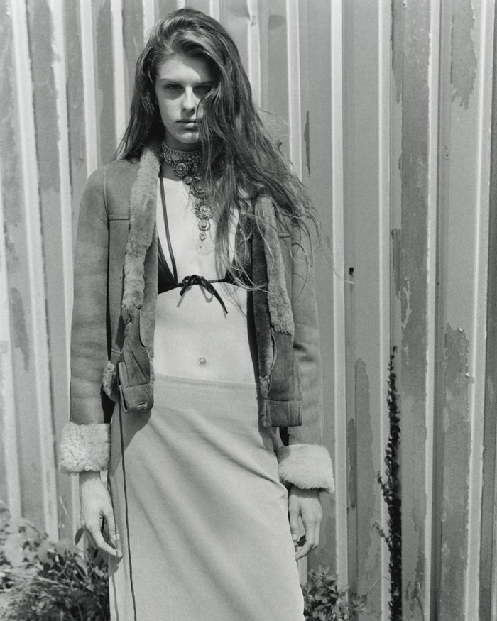 Monochrome fashion editorial portrait of a woman wearing an open shearling jacket and layered necklace against corrugated metal.