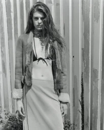 Monochrome fashion editorial portrait of a woman wearing an open shearling jacket and layered necklace against corrugated metal.