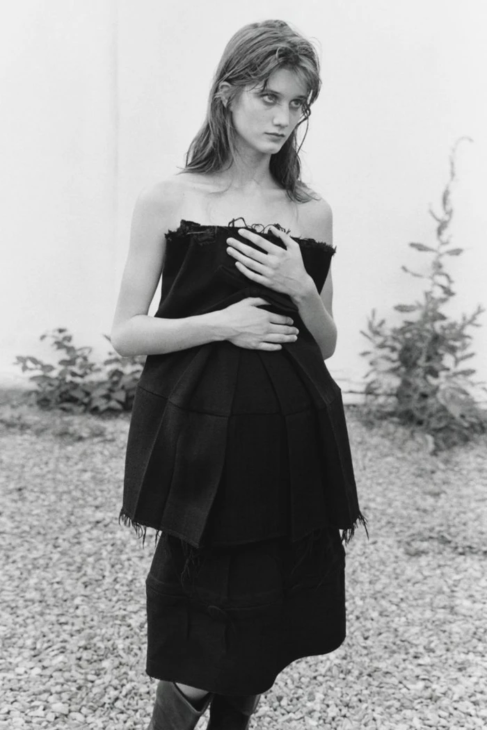 Black and white portrait of a female model wearing a structured dark strapless top against an outdoor wall.