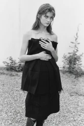 Black and white portrait of a female model wearing a structured dark strapless top against an outdoor wall.