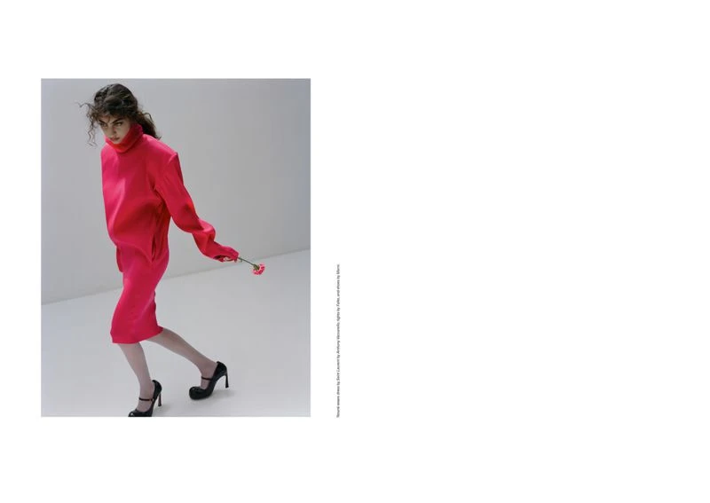 A stylish woman in a pink dress and black heels poses in a bright studio, holding a pink flower, for a fashion editorial photograph.