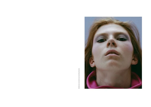 Close-up portrait of a fair-skinned adult female with auburn hair, wearing makeup and a pink collar, set against a light blue backdrop.