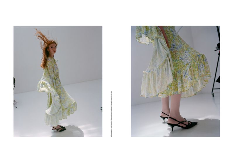 A woman wearing a floral dress poses in a bright photo studio, showcasing a full-body fashion shot with her auburn hair flying in the air.