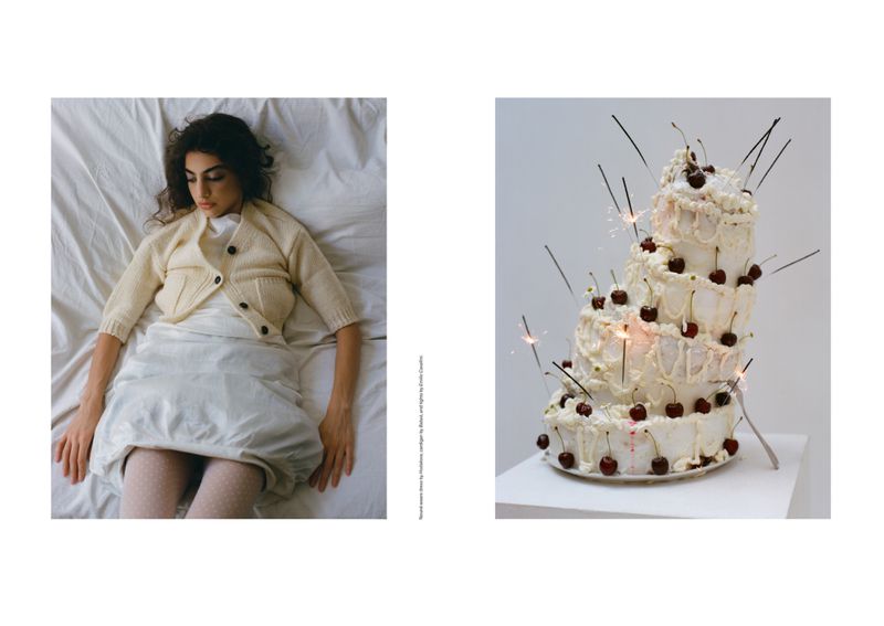 Adult female laying on a bed with a cake decorated with cherries and lit sparklers on a white pedestal against a white background.