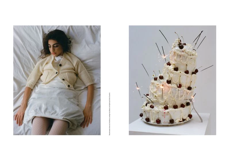 Adult female laying on a bed with a cake decorated with cherries and lit sparklers on a white pedestal against a white background.