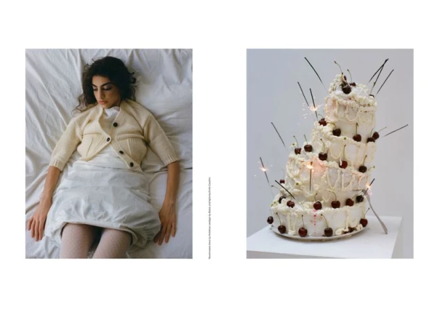 Adult female laying on a bed with a cake decorated with cherries and lit sparklers on a white pedestal against a white background.