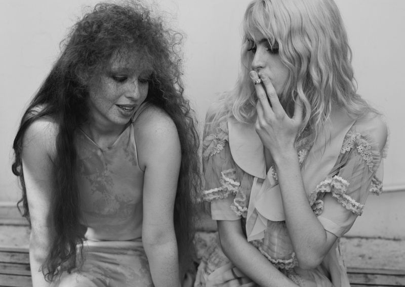 Two young women sit indoors, one smoking, in a monochrome portrait. Both wear vintage-style dresses and have distinctive hairstyles.