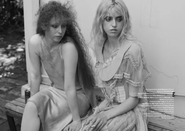 Two young female models seated together on an outdoor bench in a black and white editorial photograph.