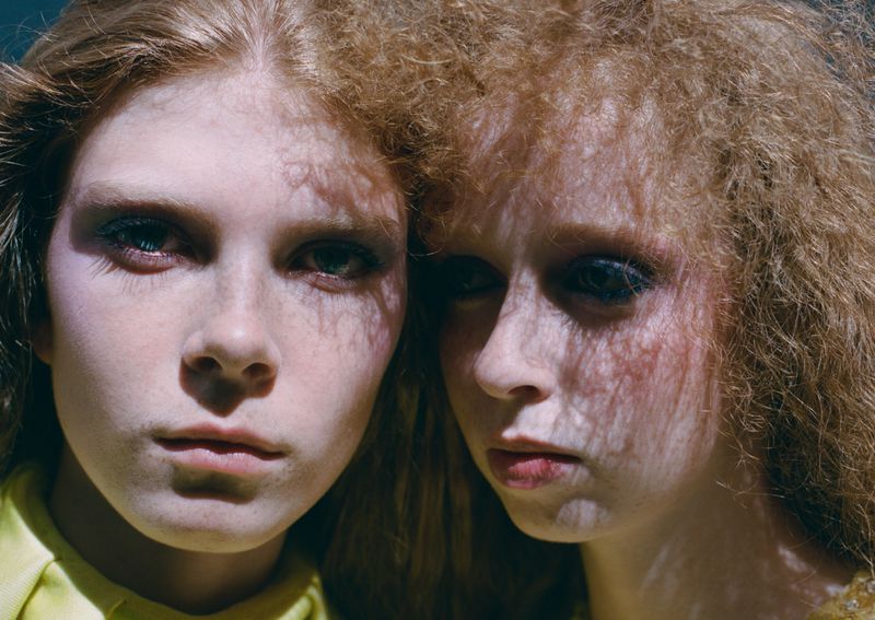 Close-up portrait of two young female models with textured red and blonde hair and dappled shadow patterns across their faces.