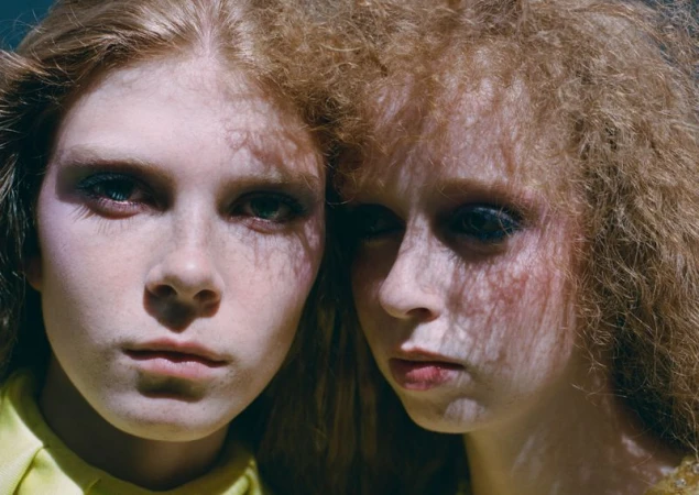 Close-up portrait of two young female models with textured red and blonde hair and dappled shadow patterns across their faces.