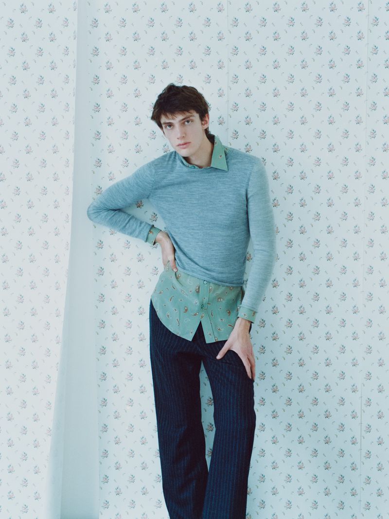 Man in blue sweater poses against floral wallpaper. He wears a green shirt and pinstripe pants in a portrait-style fashion shot.