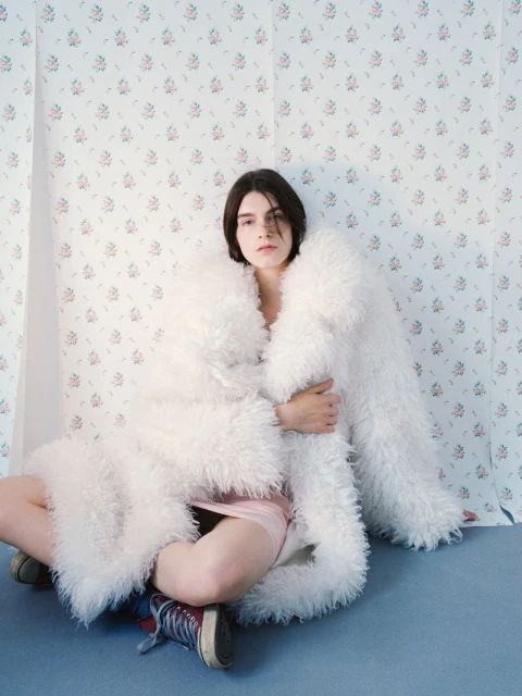 A woman in a white furry coat sits on a blue surface against a floral backdrop, wearing sneakers and a dress in a fashion pose.