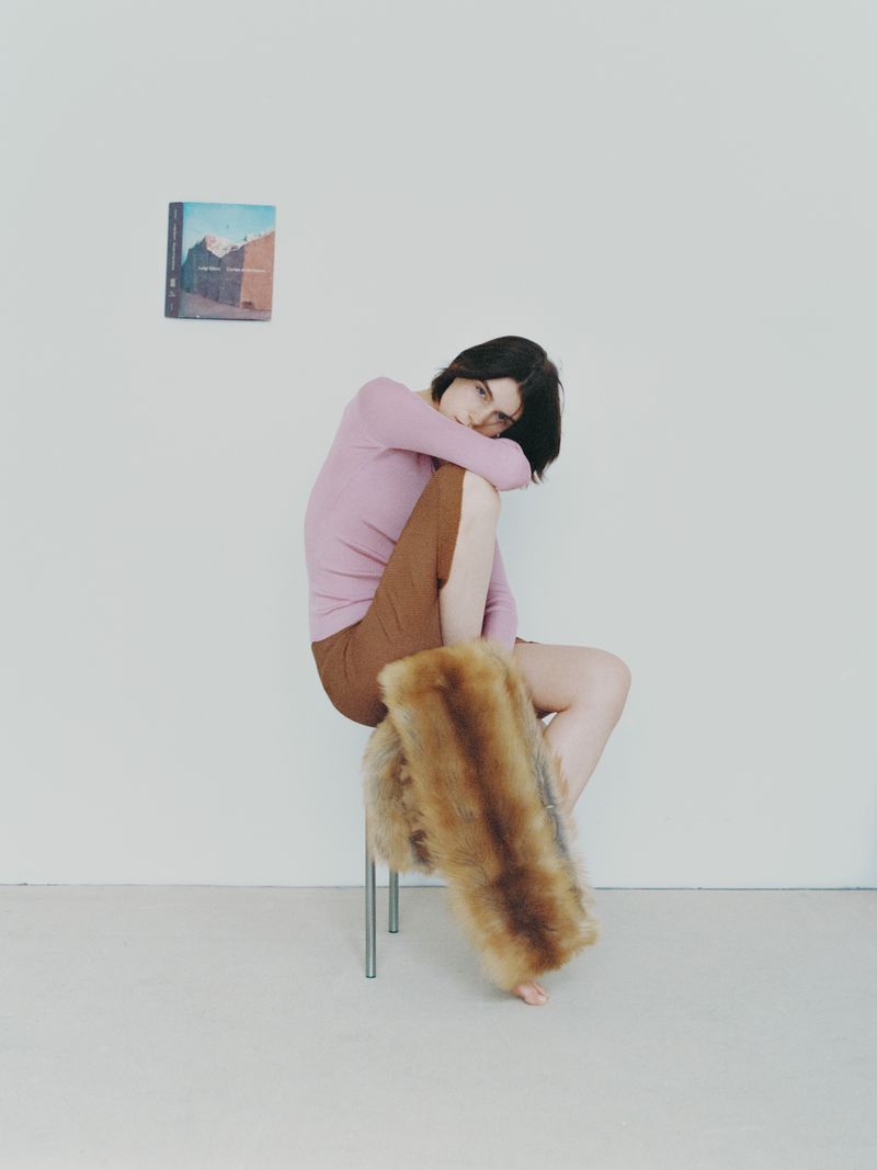 Seated young woman in pink top and brown shorts, resting arm on knee. Furry stool and painting on the wall in a bright room.