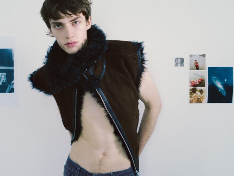 Young adult male in an unbuttoned brown leather vest with a black fur collar, posing indoors against a white wall with artwork.