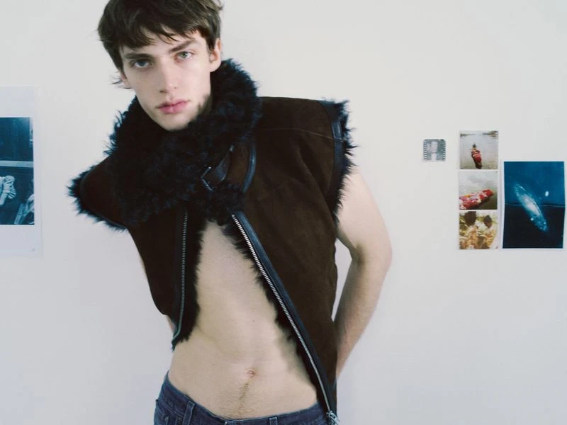Young adult male in an unbuttoned brown leather vest with a black fur collar, posing indoors against a white wall with artwork.