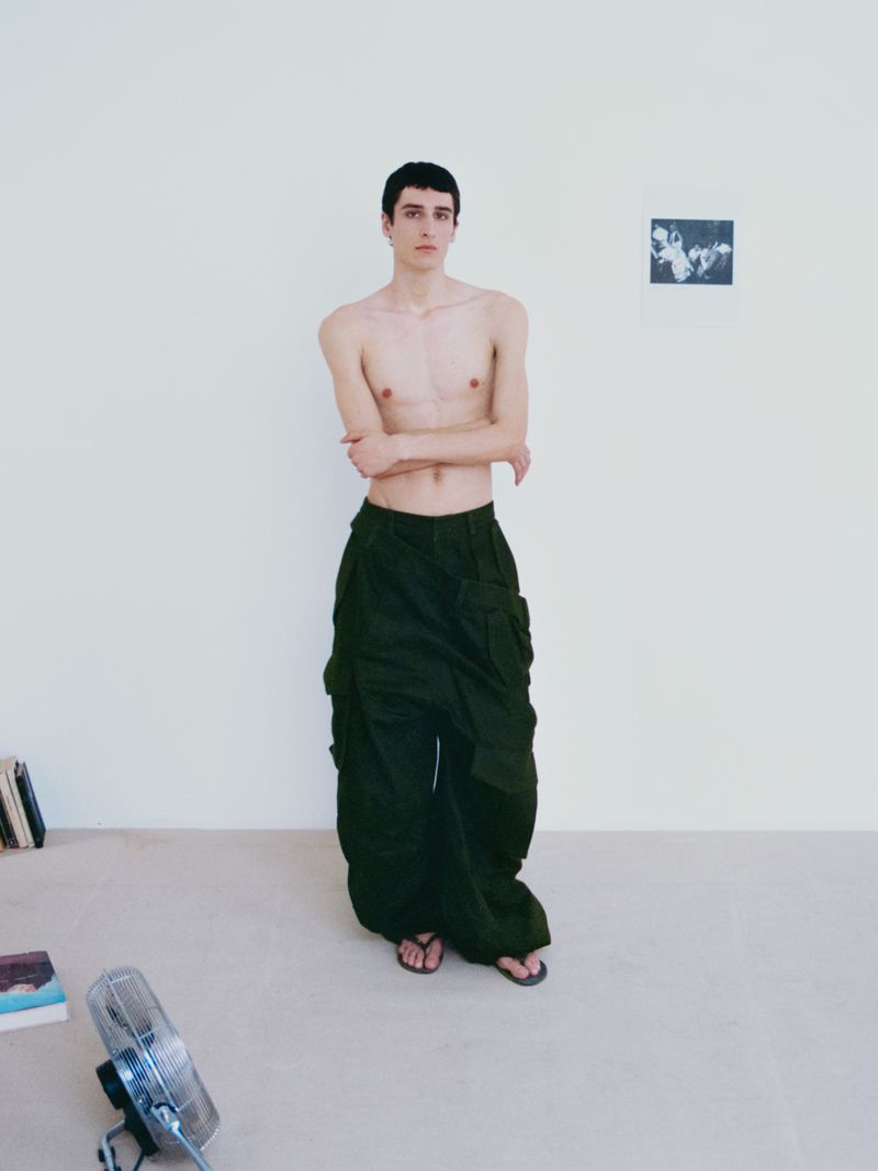 A slender, fair-skinned man stands in olive cargo pants and sandals indoors with minimalist decor, lit softly, in a full shot.