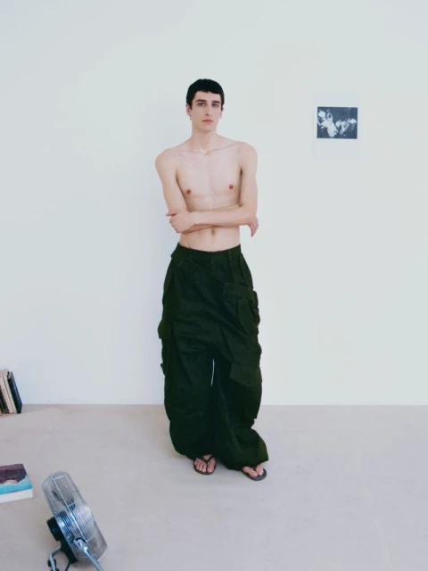 A slender, fair-skinned man stands in olive cargo pants and sandals indoors with minimalist decor, lit softly, in a full shot.
