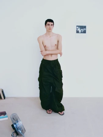 A slender, fair-skinned man stands in olive cargo pants and sandals indoors with minimalist decor, lit softly, in a full shot.