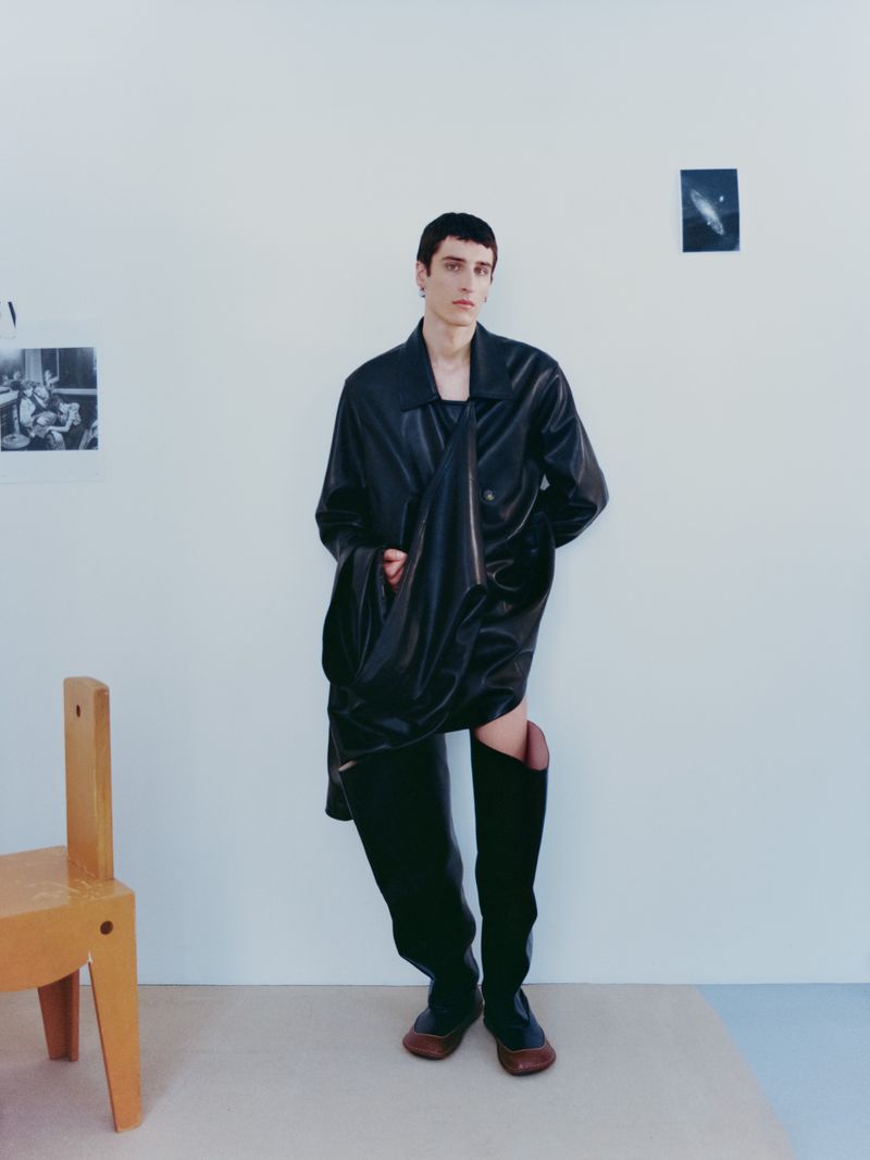 Model in black leather coat and tall boots posing against a plain wall with small framed photos