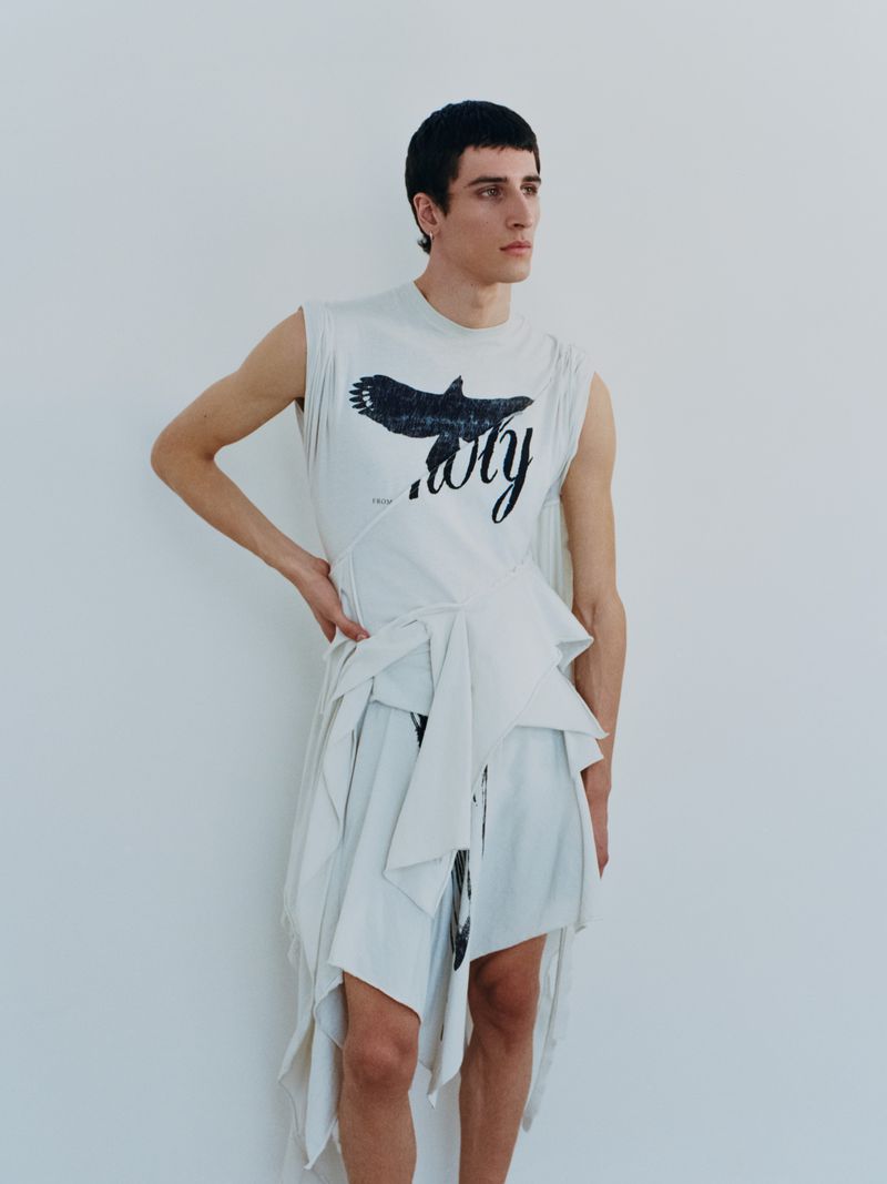 Model wearing a white sleeveless top with a bird graphic and wrapped skirt against a plain wall