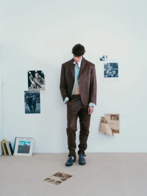 Adult male poses in a brown suit jacket and trousers against a white wall with posters, complemented by scattered newspapers and books on the floor.