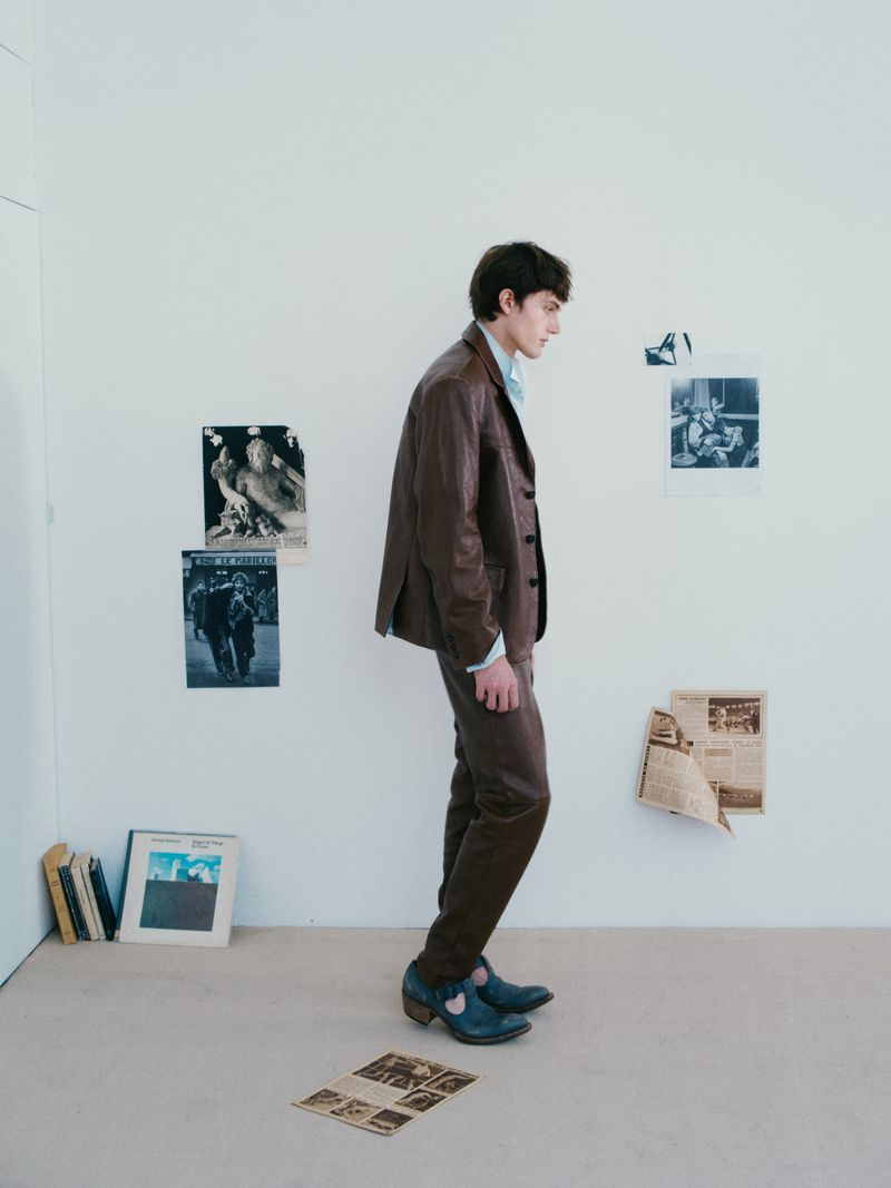 A young man in a brown suit poses in a minimalist room with posters and newspapers on the wall, creating an editorial fashion image.