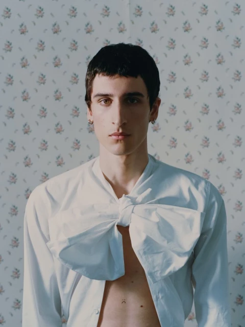 Portrait of a young adult man wearing a white shirt with a large bow on the chest, posing against a floral wallpaper backdrop indoors.