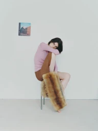 Model wearing pink knit sweater and brown skirt sitting on a stool against a plain white wall