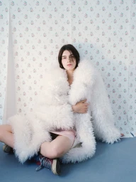 A model wearing an oversized white shaggy coat sits cross-legged on a pale blue floor in front of floral wallpaper.