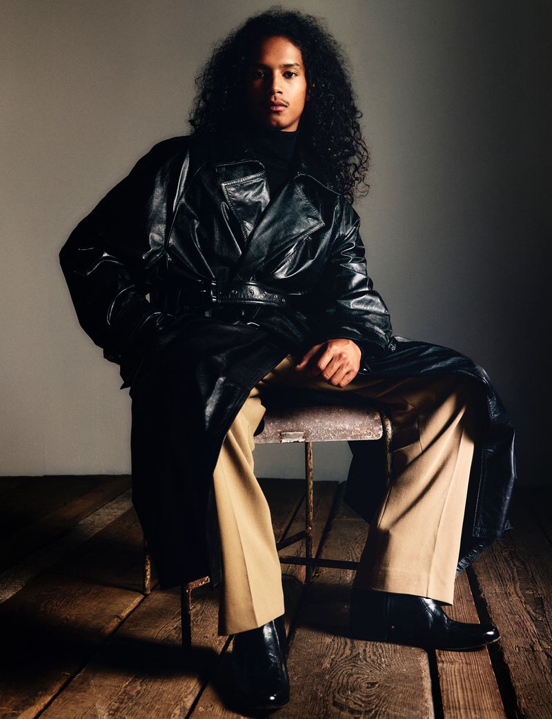Stylish adult male with curly hair sits on a wood stool, wearing a black vinyl trench coat, khaki trousers, and black boots in a studio.