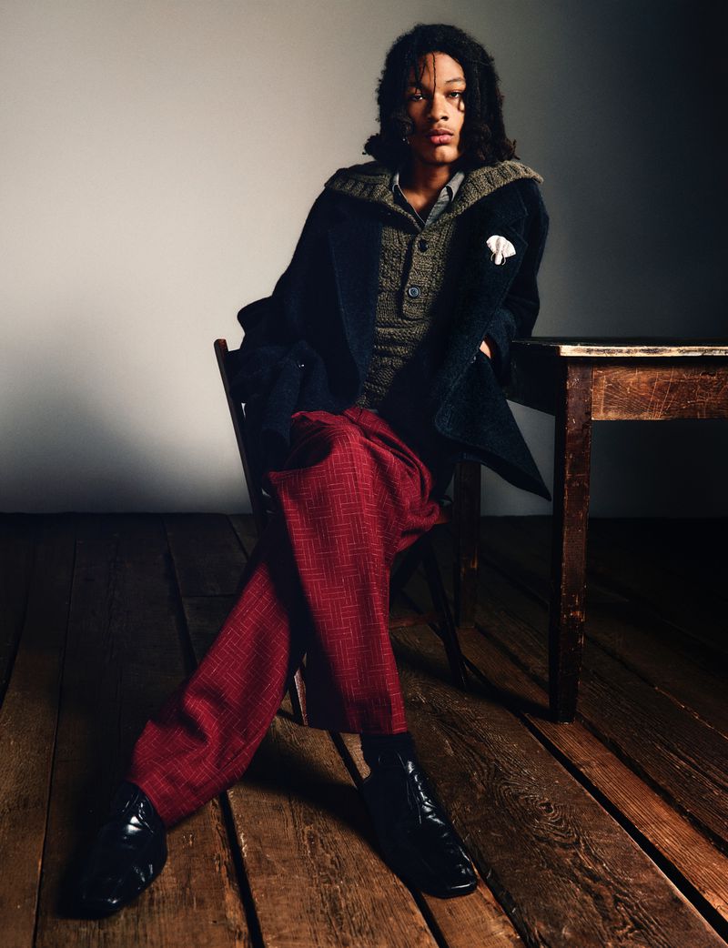 Person with long dark hair sitting on a wooden chair in red trousers and black jacket, with a wooden table next to them.