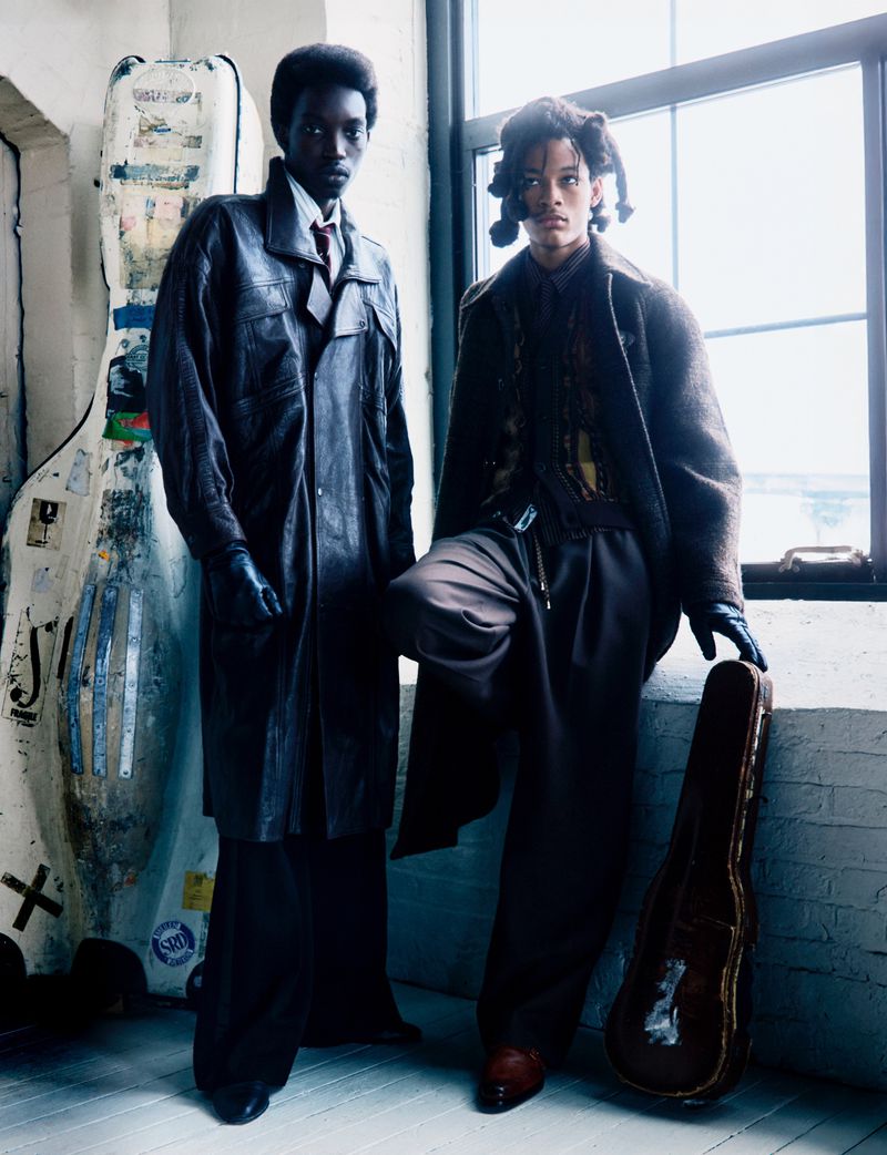 Two stylish adult models in oversized coats pose in front of a window, showcasing a fashion editorial shot with a cello case visible.