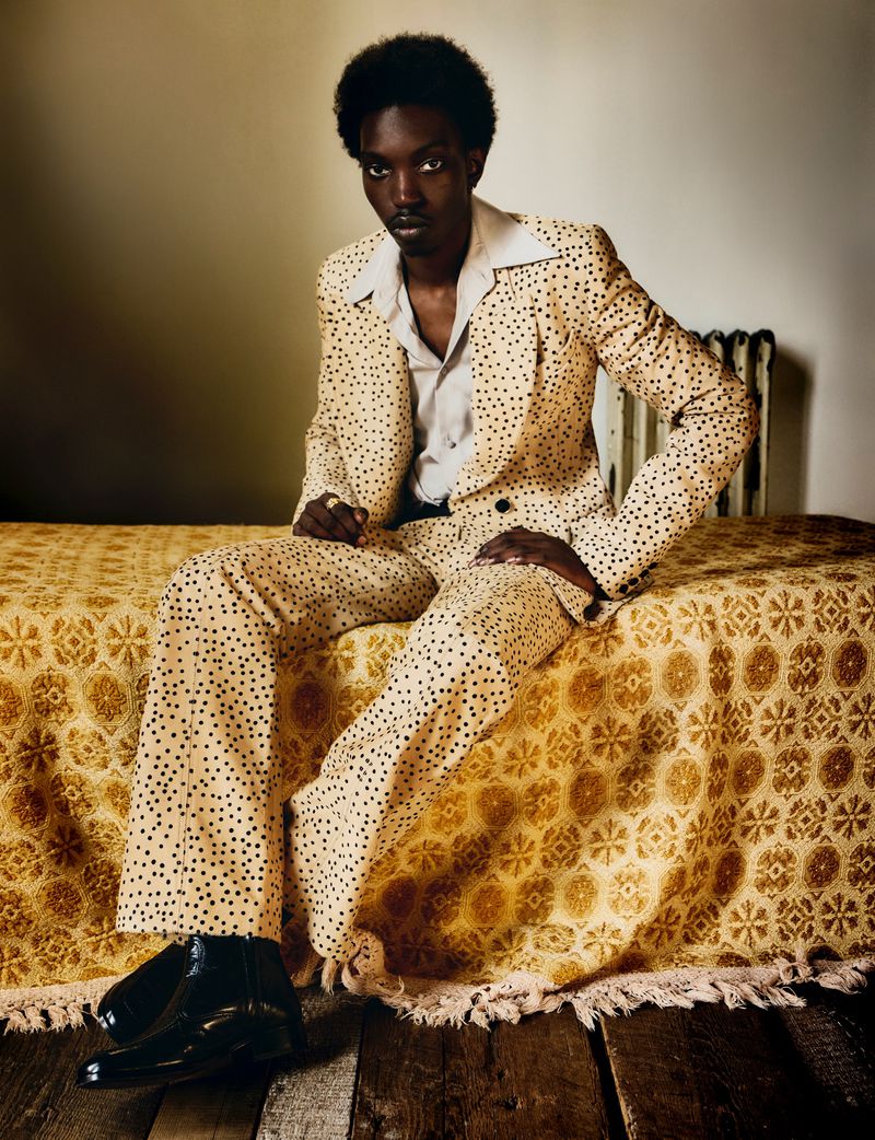 Portrait of an African-American man in a polka-dot suit, sitting on a patterned bed with a radiator in the background.