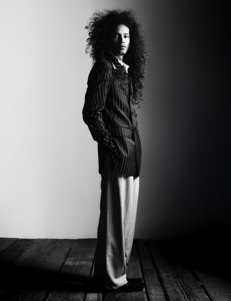 Full body portrait of a stylish adult male with curly hair in a pinstripe suit posing in a studio with a gradient light effect.