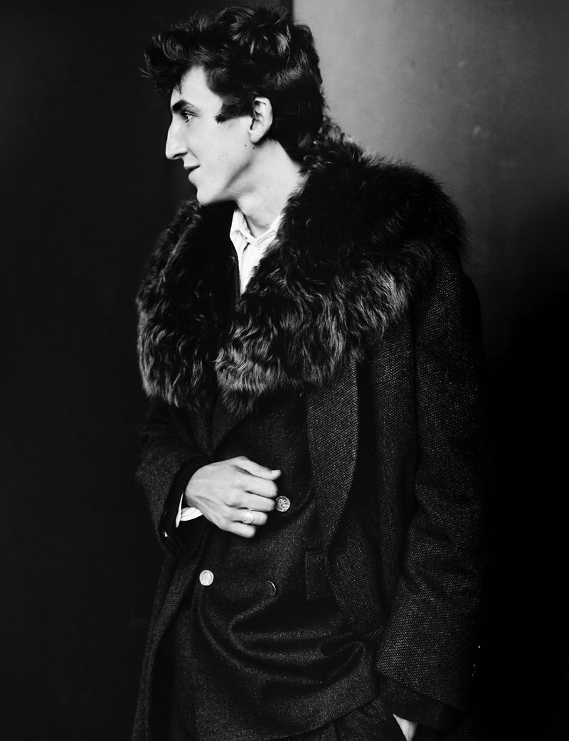 Black and white portrait of an adult male with dark hair wearing a tweed suit and a fur-collared overcoat indoors.