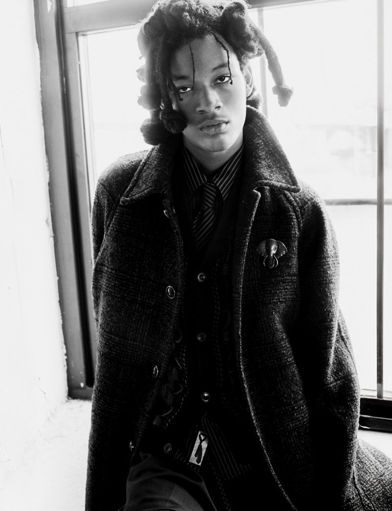 Black and white portrait of a young adult wearing a patterned overcoat, vest, and tie, posing by a window with a serious expression.