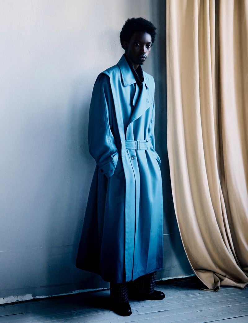 Young black man wearing long blue coat standing in front of wall and curtains, fashionable and formal, full length studio portrait.