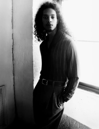 Monochrome portrait of a man with long curly hair wearing a striped collared shirt and tailored trousers standing beside a window.