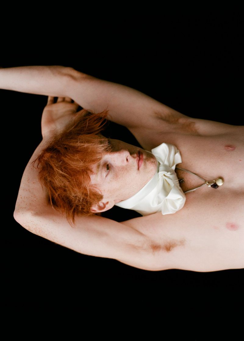 Topless male portrait with light skin and red hair lying against black background wearing a knotted, draped fabric scarf around his neck.