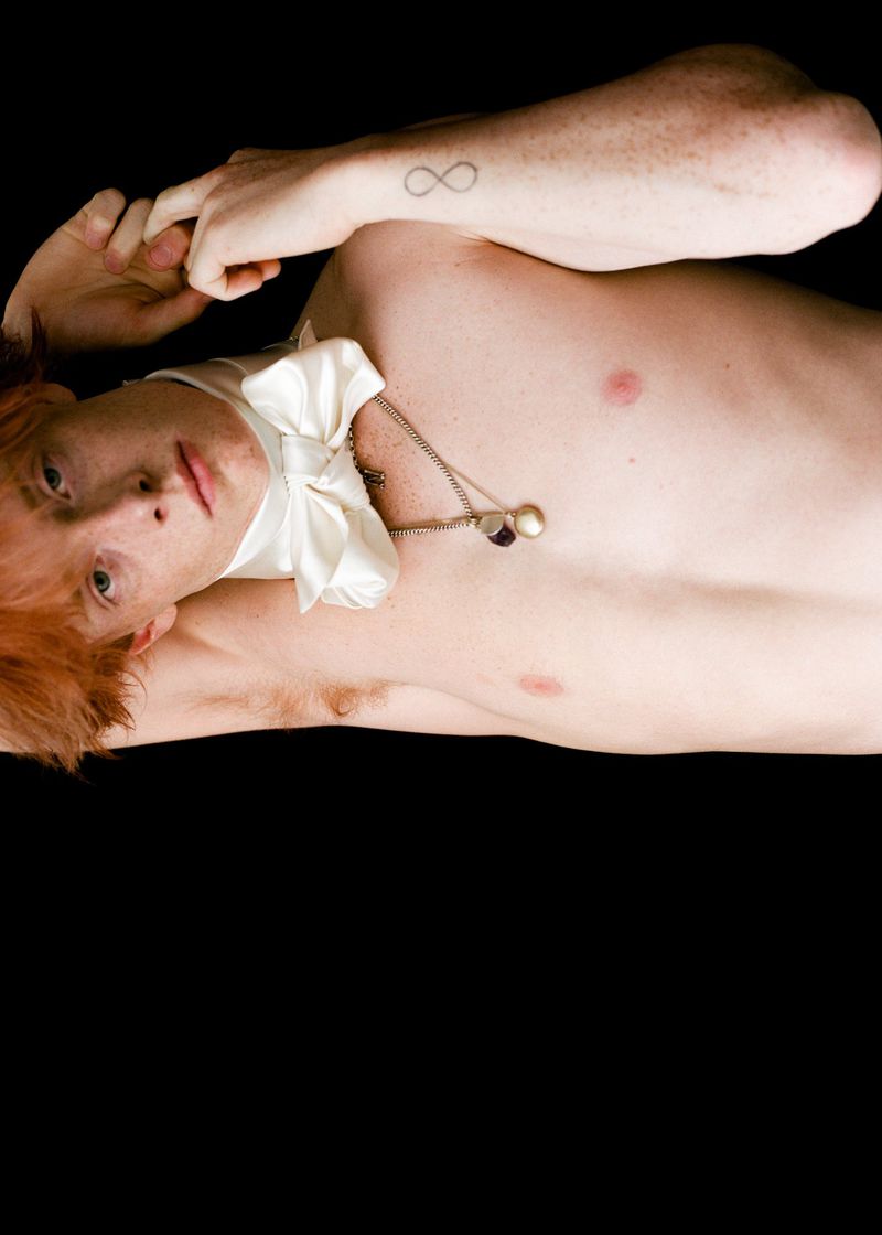 Red-haired person lying on black background wearing a white satin bow tie and necklace. Infinity tattoo on arm. Studio lighting.