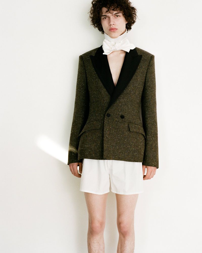 Young adult male fashion model poses in an olive green double-breasted blazer, white shorts, and a white bow against a white wall.