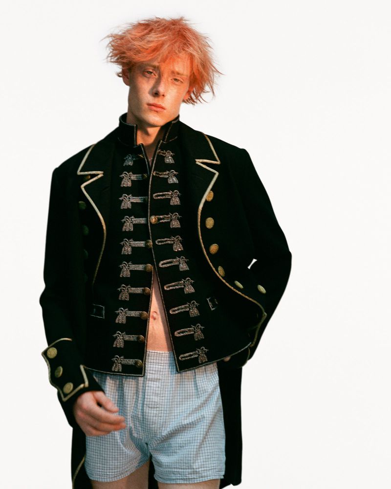 Portrait of a young adult male model with ginger hair wearing a black, decorated military-style jacket and boxer shorts in a photo studio.