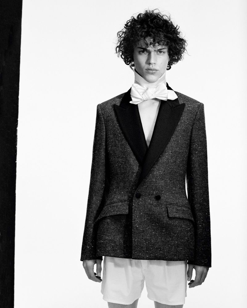 Monochrome portrait of a male model with curly hair wearing a tuxedo jacket, white shirt, and bow tie against a bright white backdrop.