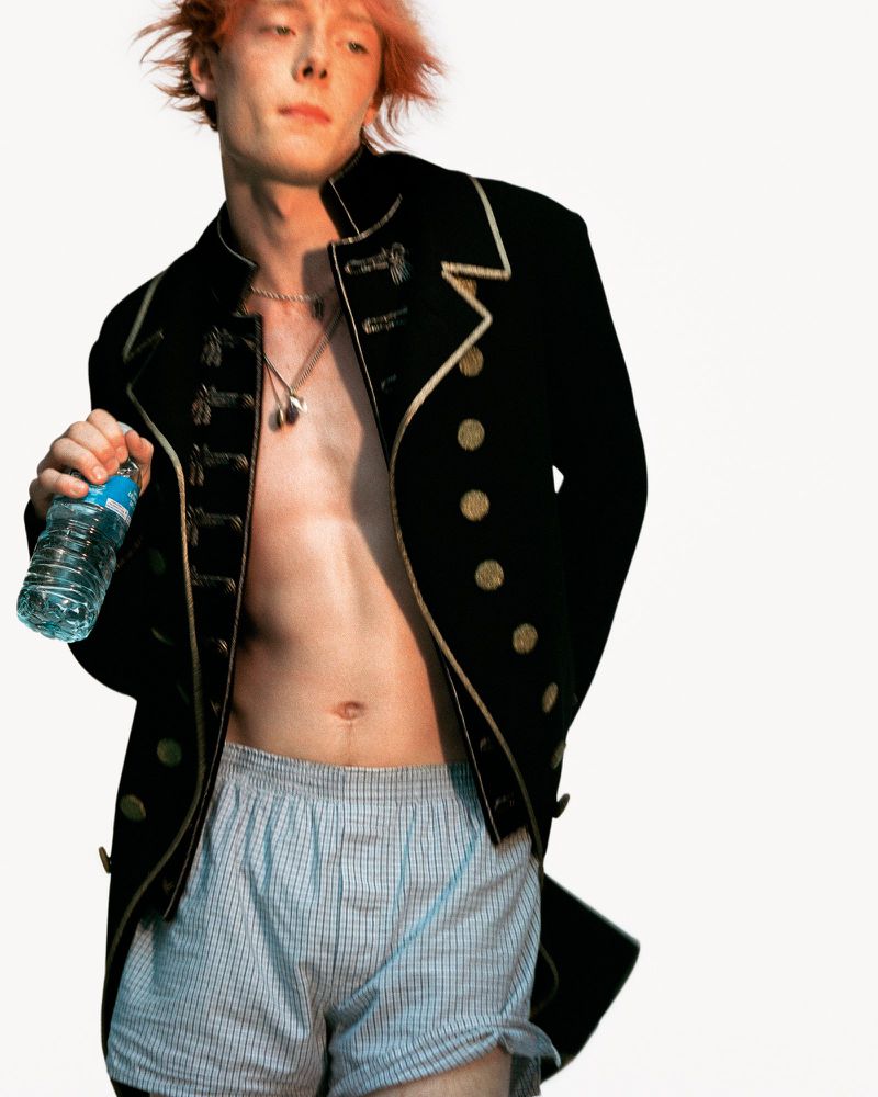 Young adult male with pink hair poses in unbuttoned jacket, boxer shorts, holding a water bottle against a white background.