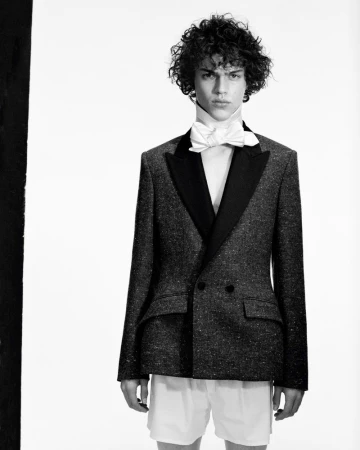 Model with curly dark hair wearing a tuxedo jacket and bow tie against a white background for Harper's Bazaar France Homme